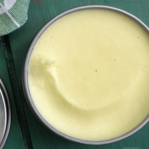 Puede incluir: Primer plano de una lata redonda de plata llena de un bálsamo amarillo pálido. El bálsamo tiene una textura suave y cremosa.