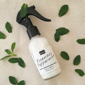 May include: A clear glass bottle with a black spray nozzle filled with a white liquid. The bottle has a white label with the text "SOW Rosemary Peppermint Hair Conditioning Mist Spray Made in California Net Wt. 4.37 oz". The bottle is surrounded by green mint leaves on a white fabric background.