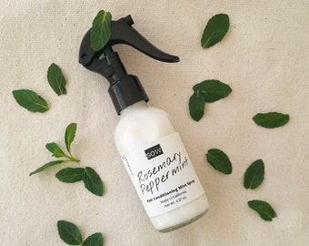 May include: A clear glass bottle with a black spray nozzle filled with a white liquid. The bottle has a white label with the text "SOW Rosemary Peppermint Hair Conditioning Mist Spray Made in California Net Wt. 4.37 oz". The bottle is surrounded by green mint leaves on a white fabric background.