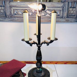 May include: An ornate, dark bronze candelabra-style lamp with three cream-colored candle-shaped bulbs. The lamp has a decorative base and arms, and a clear glass light bulb. A stack of books with red covers is visible.