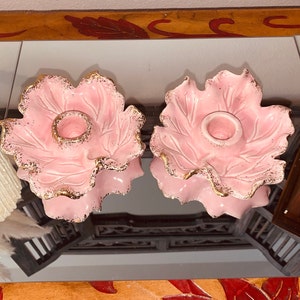 May include: Two pink ceramic candle holders in a leaf shape with gold trim. The candle holders have a circular opening for the candle and are displayed on a reflective surface. The overall theme is vintage home decor.
