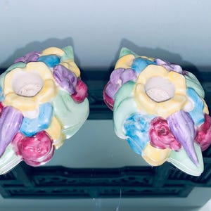 May include: Two floral-shaped candle holders. Each holder is a cluster of colorful flowers in yellow, blue, purple, pink, and green. The candle holders are made of ceramic and have a glossy finish.