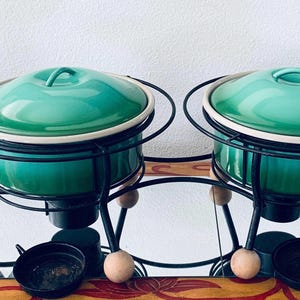 以下が含まれることがあります： 蓋付きのティール色のフォンデュ鍋2つ。黒い金属製のスタンドに木製のボール型の足が付いています。鍋には白い縁があり、鏡面の上に置かれています。小さな黒いボウルが前にあります。