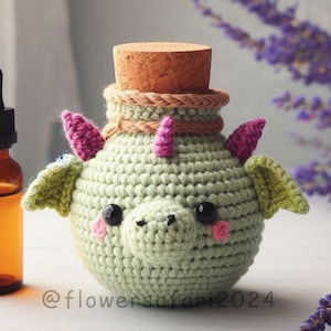 May include: A light green crocheted dragon-shaped bottle cover with pink horns and green wings, topped with a cork. A brown glass bottle with a dropper is next to the dragon. The dragon has black eyes and pink cheeks.