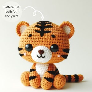 May include: A crocheted tiger plush toy with orange and black stripes, white fur on the chest and belly, and black felt eyes. The toy is sitting on a white surface.