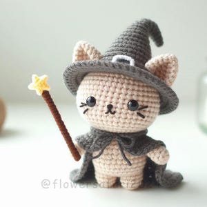 Peut inclure: Figurine de chat au crochet, portant un chapeau et une cape gris de sorcière, tenant une baguette marron avec une étoile jaune. Le chat a des yeux et des moustaches noirs. La figurine est beige et grise.