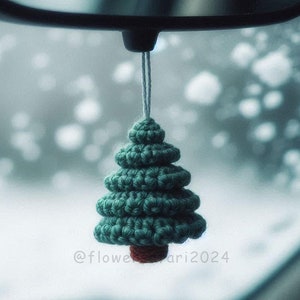 Könnte beinhalten: Ein kleiner grüner gehäkelter Weihnachtsbaum-Anhänger, der an einem Rückspiegel in einem Auto hängt. Der Baum hat eine rote Basis und ist mit weißem Garn verziert.