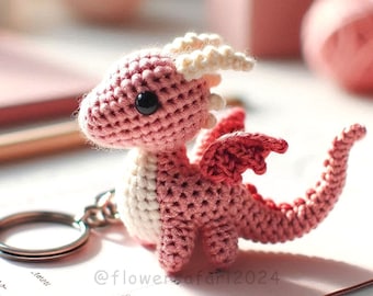 May include: A pink crocheted dragon keyring with white accents and black eyes. The dragon has a small, curled tail and wings. The keyring is attached to a silver metal ring.