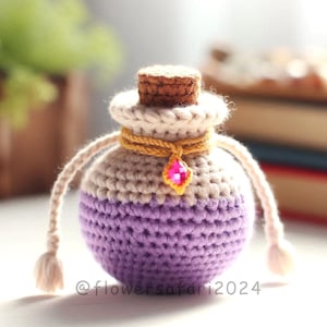 May include: A handcrafted crochet potion bottle in shades of purple and cream, adorned with a gold-colored cord and a pink gem pendant. The bottle has a cork stopper and cream-colored handles. The text "@flowersafari2024" is visible.