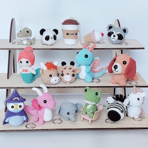 Puede incluir: Una colección de llaveros de animales y personajes hechos a crochet, que incluyen un panda, una sirena, un dragón, una jirafa, una rana, un perro, una taza de café y un unicornio. Los llaveros se muestran en un estante de madera.