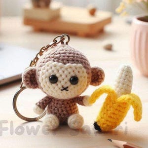 May include: Crocheted monkey keychain with a matching banana. The monkey is brown and cream-colored with black eyes and a stitched mouth. The banana is yellow with a black tip. The keychain is on a wooden surface.