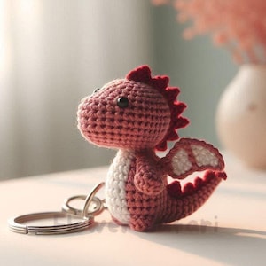 May include: A handmade crochet dragon keychain in shades of red and white. The dragon has a spiky crest, small wings, and a tail. The keychain is attached to a silver metal ring. The background is blurred, with a vase of pink flowers.