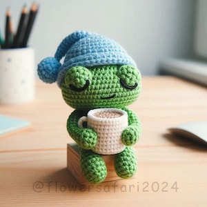 Puede incluir: Una rana de crochet verde con un gorro de dormir de crochet azul sostiene una taza blanca con un líquido marrón en su interior. La rana está sentada en un pequeño bloque de madera. El texto "@flowersafari2024" es visible en la esquina inferior derecha de la imagen.