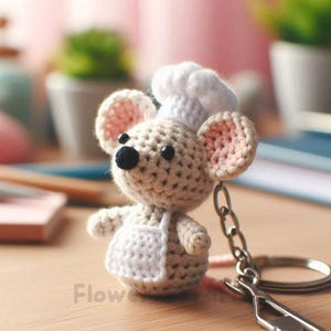 以下が含まれることがあります： コック帽とエプロンを着た、かぎ針編みのネズミのキーホルダー。ネズミはピンクの耳、黒い鼻、黒い目をしています。キーホルダーはシルバーの金属製の留め金とリングに付いています。「Flow」の文字が表面に印刷されています。