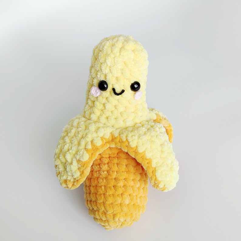May include: A crocheted banana plush toy with a smiling face and pink cheeks. The banana is yellow and has a brown stem.