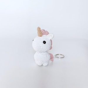 May include: White crocheted unicorn keychain with a pink mane and a gold horn. The keychain has a silver metal ring.