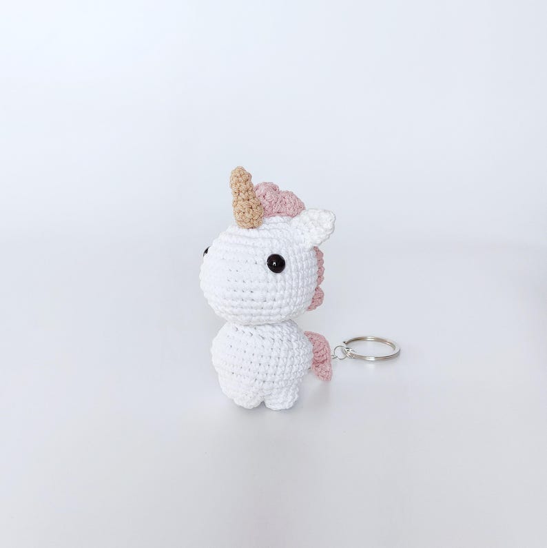 May include: White crocheted unicorn keychain with a pink mane and a gold horn. The keychain has a silver metal ring.