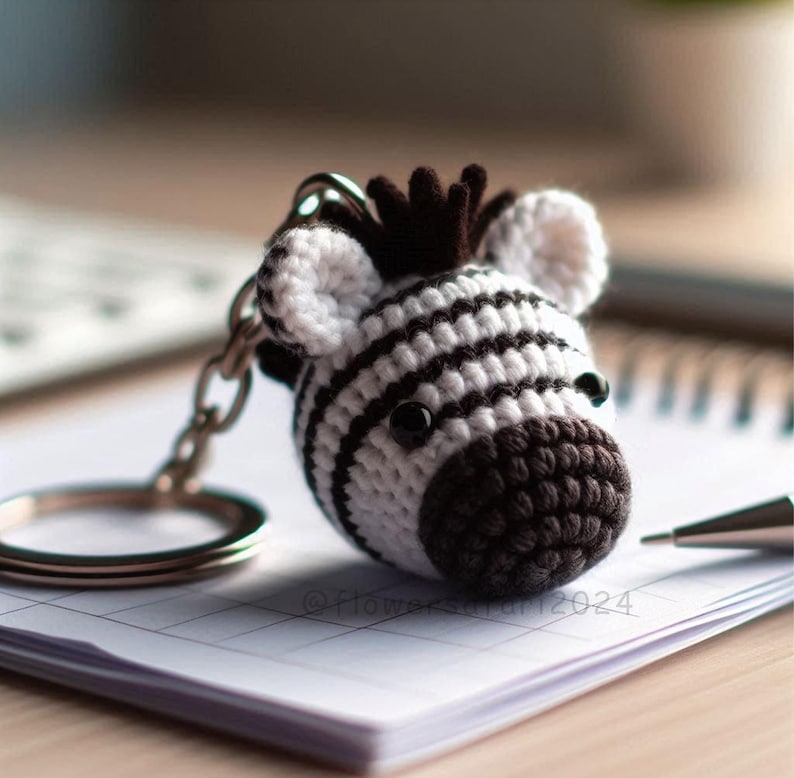 Puede incluir: Un llavero de cebra tejido a crochet con rayas blancas y negras. La cebra tiene la nariz y los ojos negros. El llavero est&aacute; unido a un anillo de llave plateado.