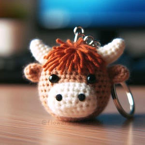 May include: A crocheted Highland cow keychain. The cow has a brown body, white horns, and a white muzzle. The keychain features a silver-colored ring and chain. The cow's hair is a reddish-brown color. The eyes are black.