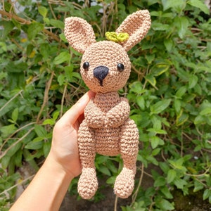 May include: A crocheted brown bunny rabbit with black eyes and a green leaf on its head. The bunny is sitting with its paws crossed.