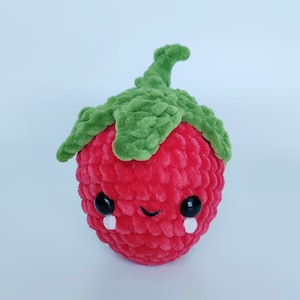 May include: A red and green crocheted strawberry plush toy with a cute, sad face. The strawberry has black eyes, a white smile, and white teardrop shapes.
