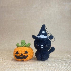 May include: Crocheted Halloween decorations featuring an orange pumpkin with a carved face and a black cat wearing a witch hat. The cat has a keychain attached. The pumpkin has a green stem. The background is a neutral tan color.