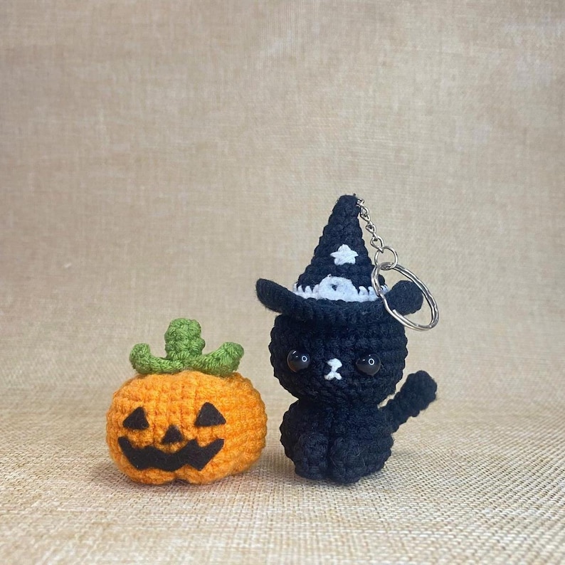 May include: Crocheted Halloween decorations featuring an orange pumpkin with a carved face and a black cat wearing a witch hat. The cat has a keychain attached. The pumpkin has a green stem. The background is a neutral tan color.