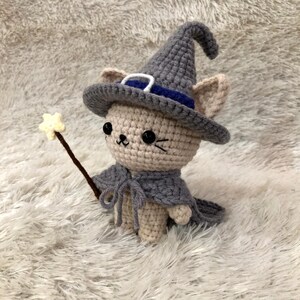 May include: A crocheted cat doll wearing a gray witch hat with a blue band, a gray cape, and holding a star-tipped wand. The cat has black eyes and a small, smiling mouth. The doll is made of beige and gray yarn.