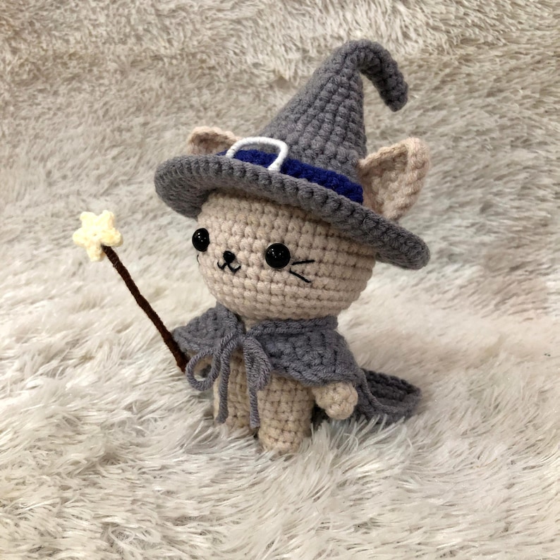 May include: A crocheted cat doll wearing a gray witch hat with a blue band, a gray cape, and holding a star-tipped wand. The cat has black eyes and a small, smiling mouth. The doll is made of beige and gray yarn.