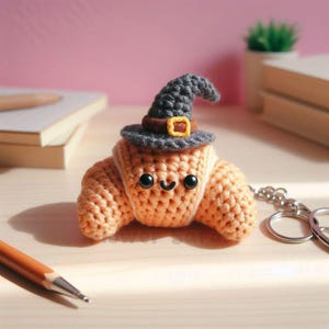 May include: A crocheted orange croissant keychain with a smiling face and a gray witch hat with a brown band and gold buckle. The keychain is on a silver chain. The background is a light pink wall and a wooden surface.