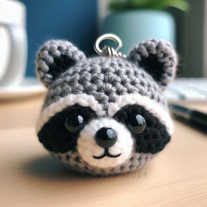 May include: A crocheted raccoon keychain with black eyes, a black nose, and a white mouth. The raccoon is gray with a white stripe around its face.