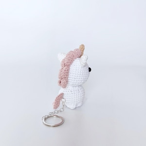 May include: White crocheted unicorn keychain with a pink mane and a gold horn. The keychain has a silver metal ring.