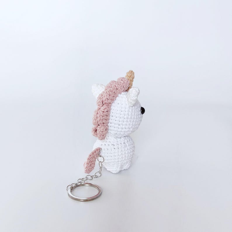 May include: White crocheted unicorn keychain with a pink mane and a gold horn. The keychain has a silver metal ring.