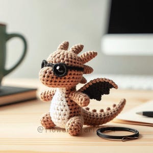 Puede incluir: Una figura de dragón de ganchillo en tonos marrones y blancos, con gafas negras. El dragón tiene alas negras y una larga cola. La figura está sobre una superficie de madera.