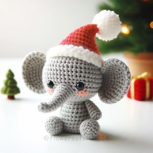 Puede incluir: Un elefante de crochet gris que lleva un gorro de Papá Noel rojo y blanco con un pompón blanco. El elefante tiene ojos negros y mejillas rosadas.