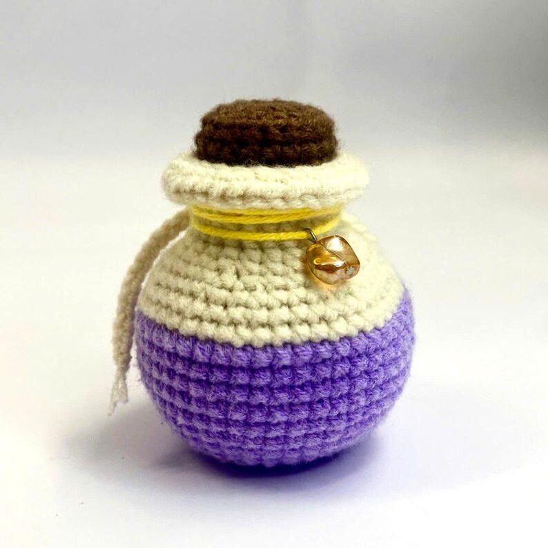 May include: A crocheted potion bottle with a brown stopper, cream-colored body, and purple base. A yellow cord wraps around the neck, and a small gold-colored bell hangs from it. The bottle has a whimsical, handmade appearance.