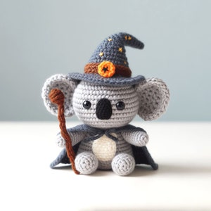May include: A gray crocheted koala wearing a blue witch hat with orange stars and a brown and orange pumpkin accent. The koala is holding a brown stick and wearing a blue cape.