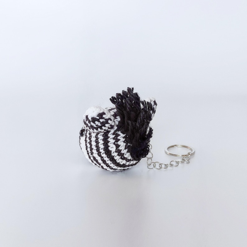 Puede incluir: Un llavero de cebra de crochet a rayas blancas y negras con una crin negra de crochet y una cadena plateada.