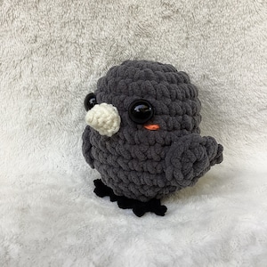 May include: A gray crocheted bird plush toy with black eyes and a white beak. The bird has a small orange mouth and is sitting on a white fluffy surface.