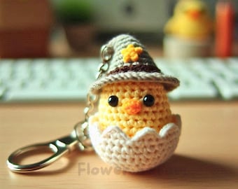 May include: A handmade crochet keychain featuring a yellow chick wearing a gray witch hat with a yellow star. The chick is inside a white crocheted eggshell. The keychain has a silver clasp and the word "Flower" is visible.