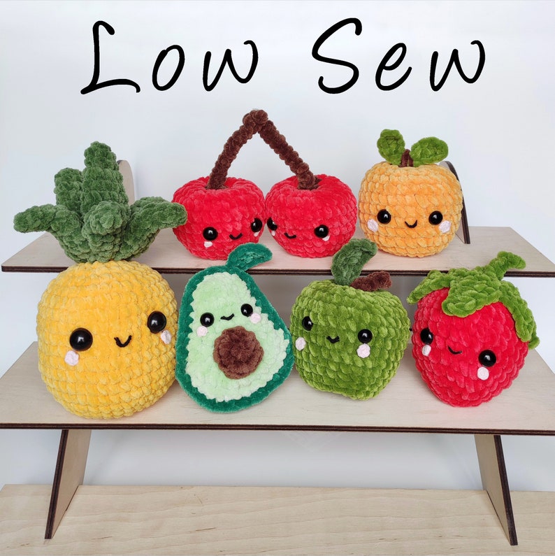 May include: Seven crocheted plush fruit toys with smiling faces. The fruits are a pineapple, two cherries, an avocado, an apple, and a strawberry. The toys are arranged on a wooden shelf with the text "Low Sew" above them.