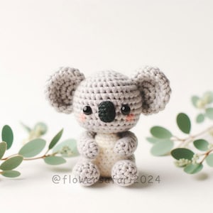May include: A crocheted koala plush toy with grey fur, black eyes, and a small black nose. The koala is sitting with its arms crossed and has a white belly.