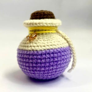 May include: A handmade, crocheted potion bottle in shades of purple, cream, and brown. The bottle has a rounded shape with a small, brown cap and is adorned with yellow yarn and a gold-colored charm. A cream-colored cord is attached.