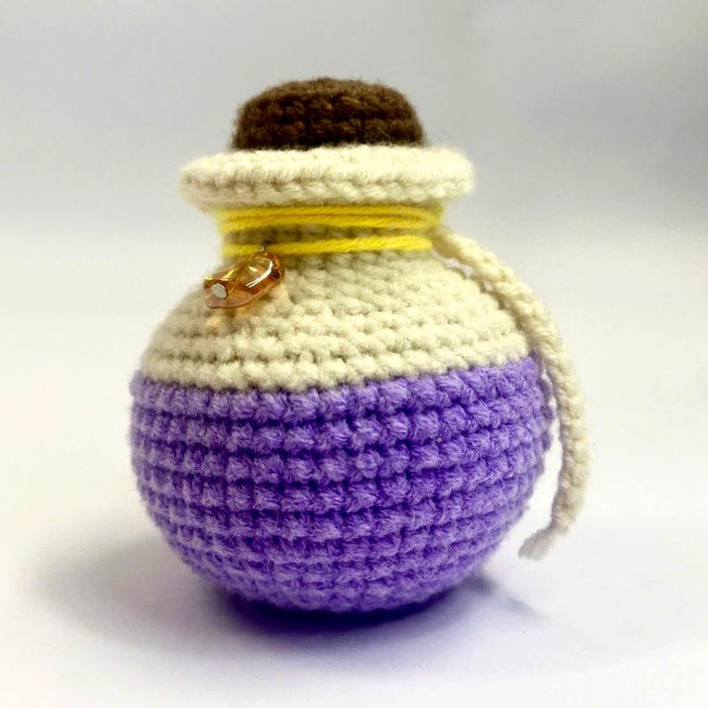 May include: A handmade, crocheted potion bottle in shades of purple, cream, and brown. The bottle has a rounded shape with a small, brown cap and is adorned with yellow yarn and a gold-colored charm. A cream-colored cord is attached.