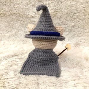 May include: A handmade crochet doll dressed as a wizard. The doll wears a gray pointed hat with a blue band, a gray cape, and has beige ears. It holds a brown wand with a star on the end. The doll is made of yarn.