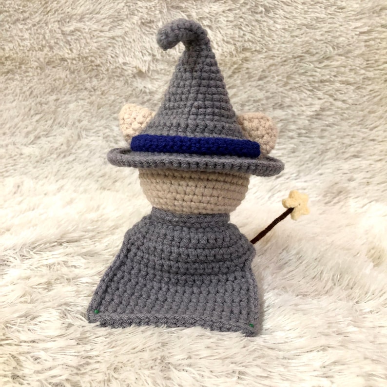 May include: A handmade crochet doll dressed as a wizard. The doll wears a gray pointed hat with a blue band, a gray cape, and has beige ears. It holds a brown wand with a star on the end. The doll is made of yarn.