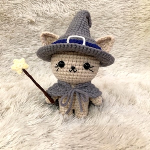 May include: A crocheted cat doll wearing a gray witch hat with a blue band and a matching cape. The cat holds a star-shaped wand. The doll is beige with black eyes and whiskers. The background is a fluffy white surface.