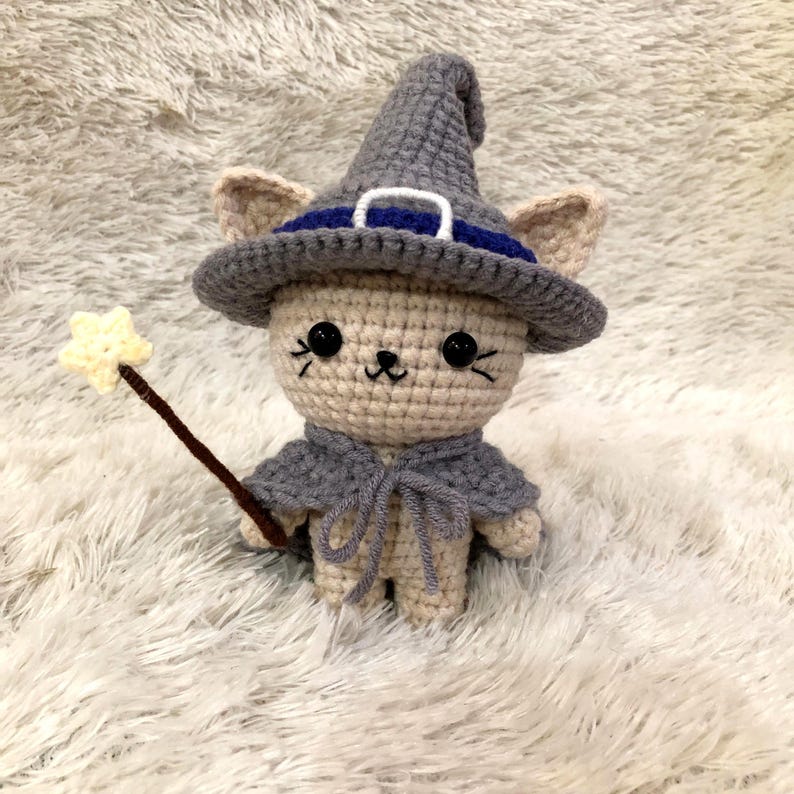 May include: A crocheted cat doll wearing a gray witch hat with a blue band and a matching cape. The cat holds a star-shaped wand. The doll is beige with black eyes and whiskers. The background is a fluffy white surface.
