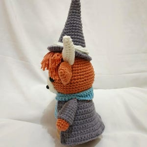 Pu&ograve; includere: Un giocattolo di peluche all'uncinetto a forma di piccolo animale che indossa un cappello a punta grigio, una sciarpa blu e un cappotto grigio.