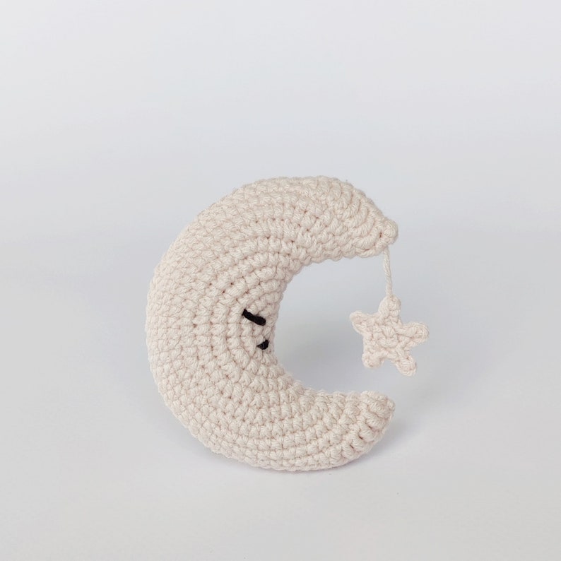 May include: A crocheted cream-colored crescent moon with a black embroidered face and a small crocheted star attached to the tip.
